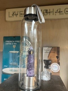 A clear glass water bottle with a stainless steel lid and a purple crystal chamber visible through the bottle's center. Well Building Connection