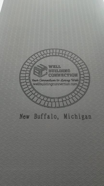 Gray yoga mat with a textured surface, displaying a logo and text indicating the Well Building Connection brand and location in New Buffalo, Michigan. Well Building Connection Exercise floor mat provides overall benefits for all fitness levels
